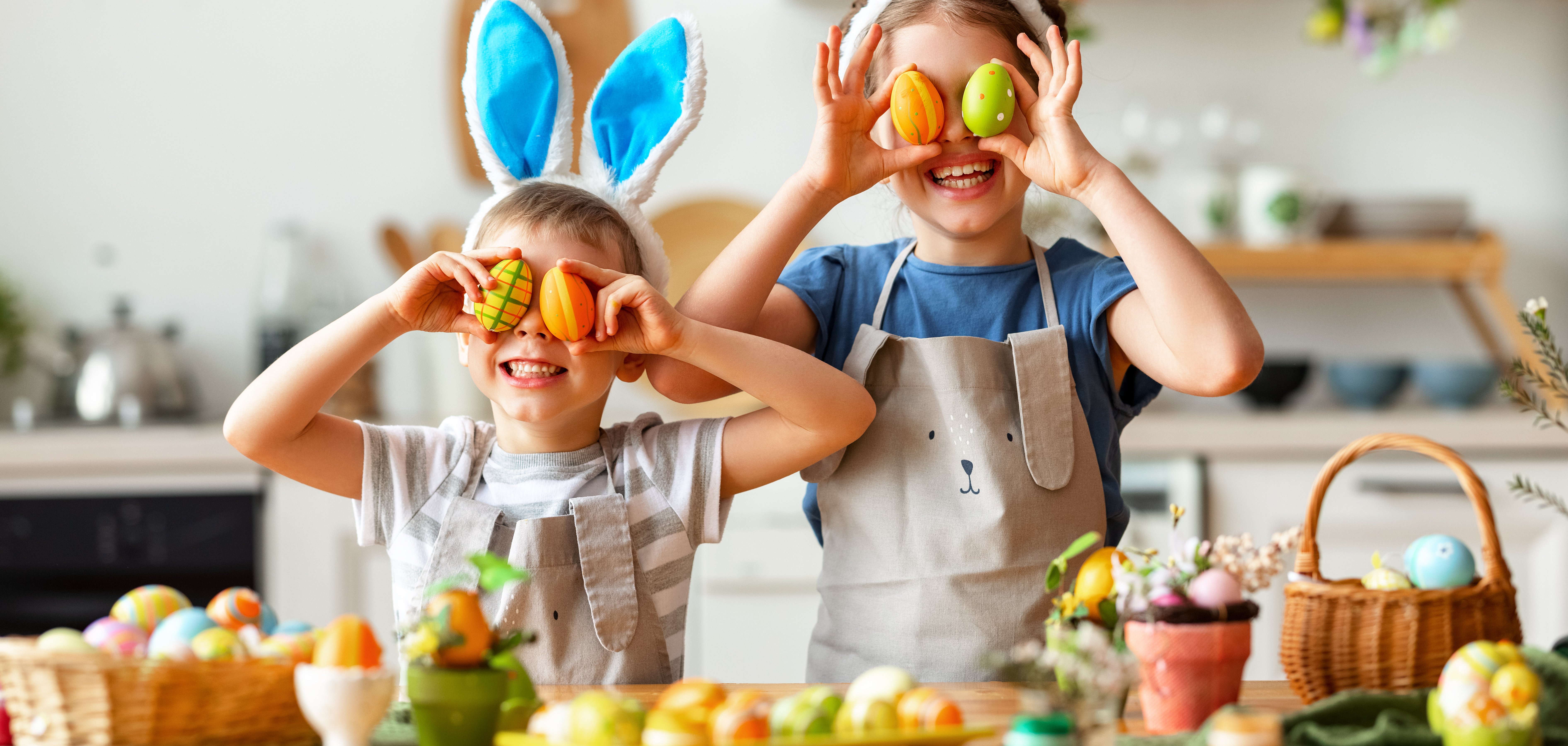 Auf einem Tisch stehen Körbe mit Ostereiern. Dahinter steht ein Junge und ein Mädchen mit Hasenohren. Sie halten sich Ostereier vor die Augen. Im Hintergrund unscharf eine Küche.