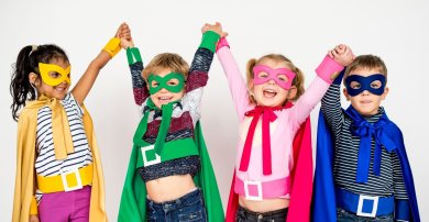 Vier mit Maske, Cape und Gürtel als Superhelden verkleidete Kinder strecken die Hände nach oben und halten sich fest. Von links nach rechts Mädchen in gelb, Junge in grün, Mädchen in rosa und Junge in blau.