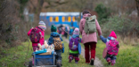 Man schaut von hinten auf eine kleine Gruppe des Waldkindergartens auf dem Weg zur Basis. Die Erzieherin hat links drei Kinder und rechts ein Kind bei sich. Alle sind winterlich gekleidet und haben Rucksäcke auf. Das linke Kind zieht einen Bollerwagen.