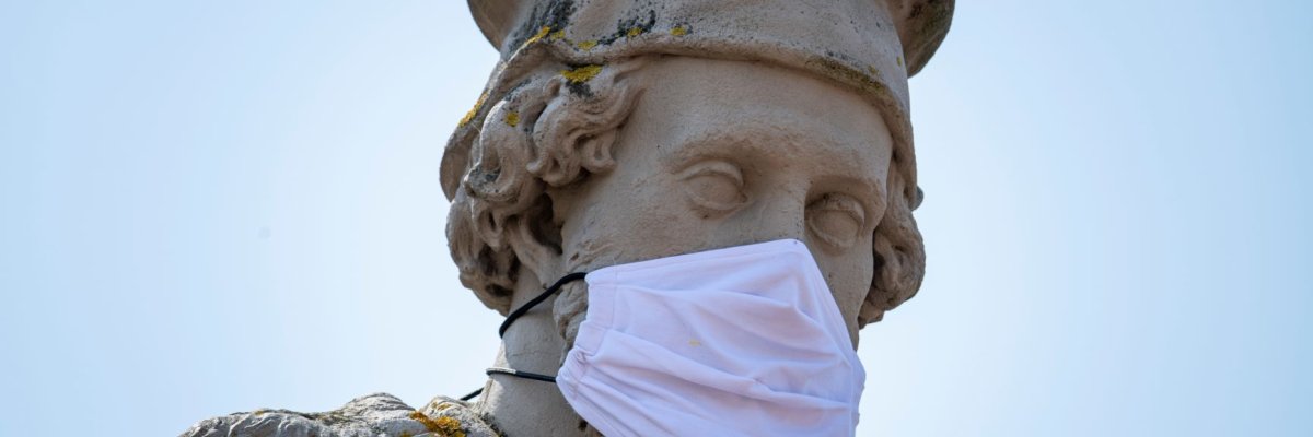 Eine Großaufnahme des Kopfs des Schöffer Denkmals Peter Schöffer trägt eine  Mundschutz aus Stoff das Bild entstand zu Beginn der Corona-Pandemie 2020