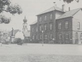 eine schwarz weiße Fotografie vom Schöfferplatz mit dem heutigen Museum als Schule noch ohne den heutigen Haupteingang und dem Denkmal davor mit der ursprünglichem Bepflanzung um den Sockel herum das Bild zeigt den Zustand um 1823 herum
