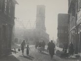 Das schwarz weiße Bild zeigt die im zweiten Weltkrieg zerstörte Innenstadt von Gernsheim ungefähr mit Blick vom heutigen Stelter Geschäft in Richtung katholischer Kirche von welcher auf dem Bild nur die Mauern und der Turm stehen und das Dach gerade wieder aufgebaut wird im Bild sieht man Menschen geschäftig durch die Magdalenenstraße gehen eine Frau läuft mir einem Kind an der Hand auf den Fotografen zu hinter ihr steht ein Fuhrwerk und rechts im Bild stehen mehrere Mütter mit Kinderwagen vor einem Haus