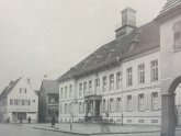 Auf dem schwarz weißen Bild sieht man das Stadthaus nach dem Wiederaufbau nach dem zweiten Weltkrieg Der Platz ist offen ohne Poller und Blumenkübel die Fenster des Stadthauses haben Holzfensterläden Menschen laufen rechts im Bild die heutige Brasserie ist noch ein Geschäft