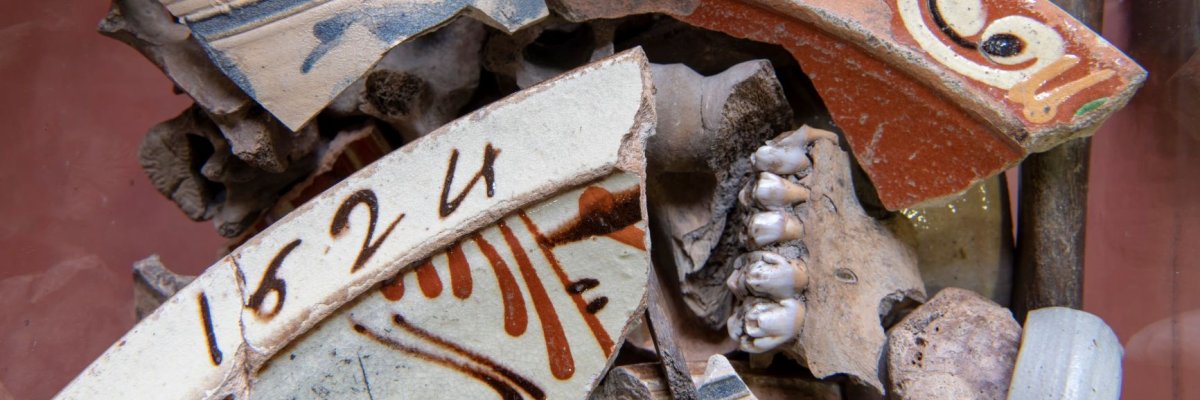 Das Bild zeigt antike Fundstücke wie Scherben von Steingut mit blauer Bemalung und Keramik mit Tonfarben sowie Knochen