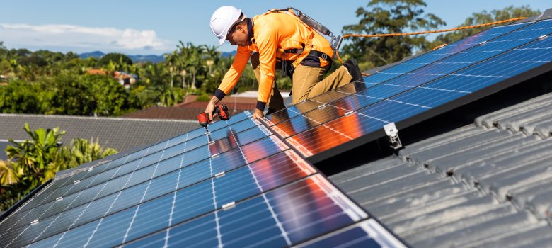 Ein Mann in orangefarbener Kleidung installiert mit einer Bohrmaschine Solarmodule auf einem Hausdach.