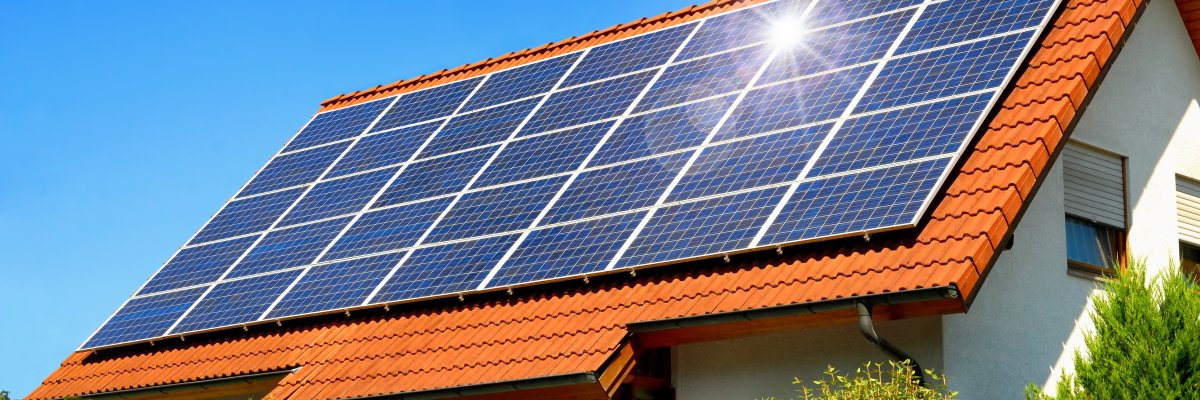 Solaranlage auf einem Hausdach unter strahlend blauen Himmel, mit der Reflektion der Sonne. Im Vordergrund befindet sich eine grüne Hecke.