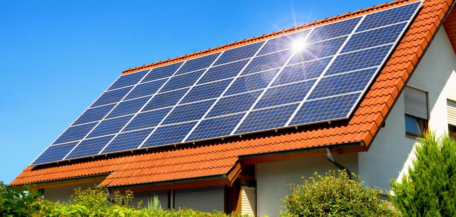 Solaranlage auf einem Hausdach unter strahlend blauen Himmel, mit der Reflektion der Sonne. Im Vordergrund befindet sich eine grüne Hecke.