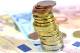 Ein Turm aus verschiedenen Euro Münzen im vorderen Zentrum des Bilds. Im Hintegrund liegen verschiedene Geldscheine und Cent Stücke.