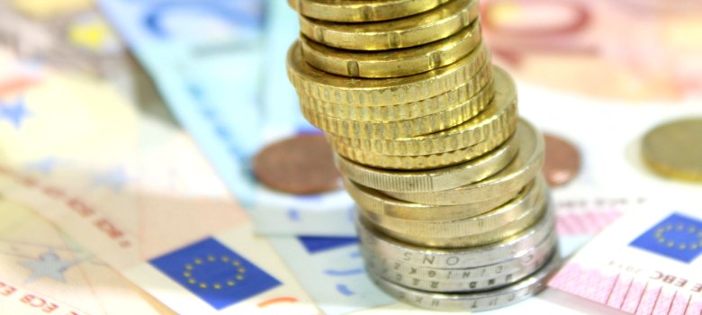 Ein Turm aus verschiedenen Euro Münzen im vorderen Zentrum des Bilds Im Hintegrund liegen verschiedene Geldscheine und Cent Stücke