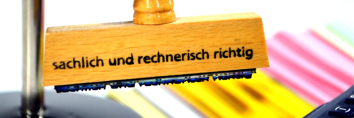 Holzstempel mit der Aufschrift sachlich rechnerisch richtig hängt in einem Stempelkarussell. Dahinter sind bunte Papierstapel. Rechts vorne in der Bildecke sind die Tasten eines Taschenrechners sichtbar.