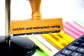 Holzstempel mit der Aufschrift sachlich rechnerisch richtig hängt in einem Stempelkarussell. Dahinter sind bunte Papierstapel. Rechts vorne in der Bildecke sind die Tasten eines Taschenrechners sichtbar.