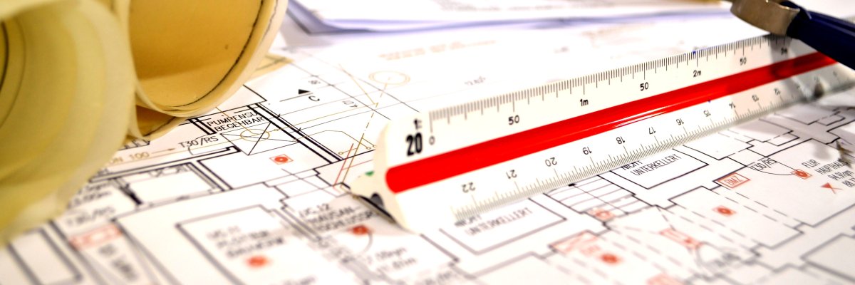 Grundrisse und Baupläne für Häuser liegen ausgebreitet und füllen das Bild aus. Darauf liegen weitere gerollte Pläne. Ein Lineal mit verschiedenen Maßeinheiten liegt im Zentrum des Bildes, darauf abgestützt, sieht man den Kopf eines Zirkels.