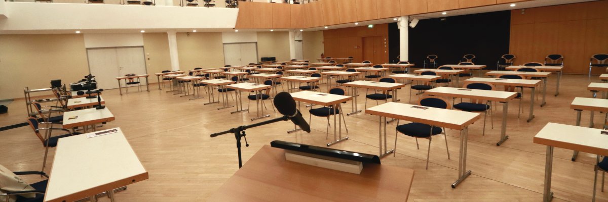 Saal der Stadthalle Gernsheim. Auf einem Parkettboden stehen einzeln in Reihe gestellte weiße Tische mit blauen Stühlen. Im Vordergrund ein Rednerpult mit Mikrofon.