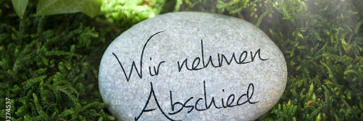 Ein heller ovaler Stein mit der Gravur "Wir nehmen Abschied" liegt auf Gras.