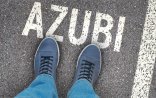 Zu sehen sind die Beine und blaue Turnschuhe einer Person, die auf einer Asphaltfläche steht. Auf dem Asphalt steht das Wort Azubi in Großbuchstaben. 