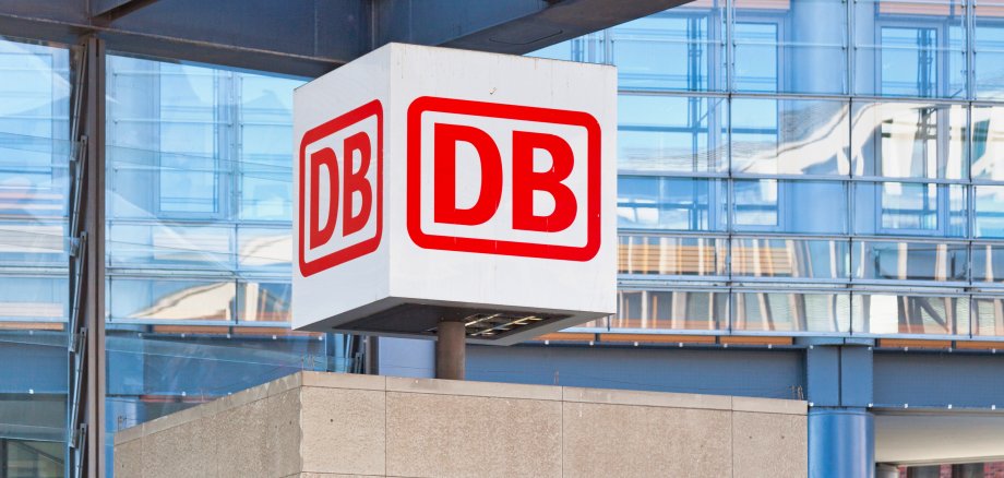 In einem Bahnhof ist das Logo der Deutschen Bahn auf einem großen, weißen Würfel zu sehen. Die Buchstaben D B sind in roter Schrift und rot umrandet. Im Hintergrund ist eine verspiegelte Glasfassade zu erkennen.