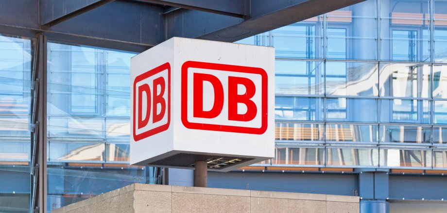 In einem Bahnhof ist das Logo der Deutschen Bahn auf einem großen, weißen Würfel zu sehen. Die Buchstaben D B sind in roter Schrift und rot umrandet. Im Hintergrund ist eine verspiegelte Glasfassade zu erkennen.