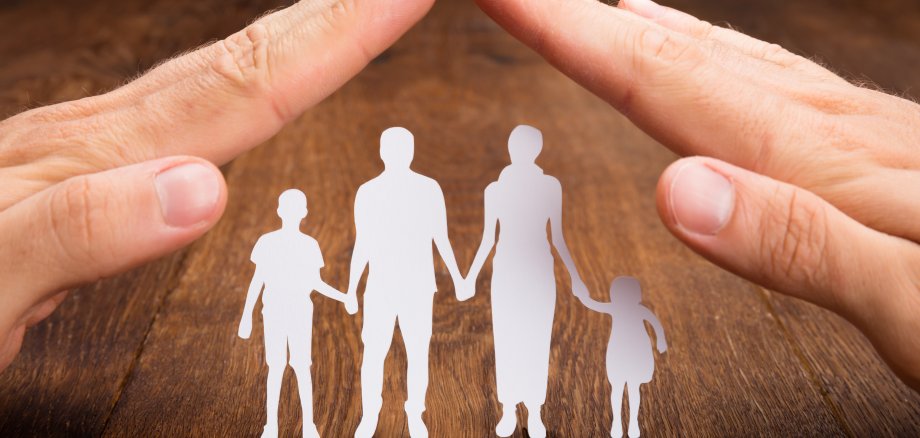 Zwei Hände werden zu schützendem Dach gehalten, unter welchen sich eine Familie befindet, die als Scherenschnitt dargestellt sind.