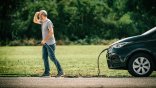 Mann hält Ladekabel in der Hand und sucht nach Ladesäule Eine männliche Person hält ein Elektroauto-Ladekabel in der Hand, welches mit einem schwarzen E-Auto verbunden ist. Das Auto ist bis zum Vorderreifen erkennbar. Der Mann hält eine Hand suchend an die Augen. Im Hintergrund ist eine Rasenfläche und ein Waldrand zu erkennen.
