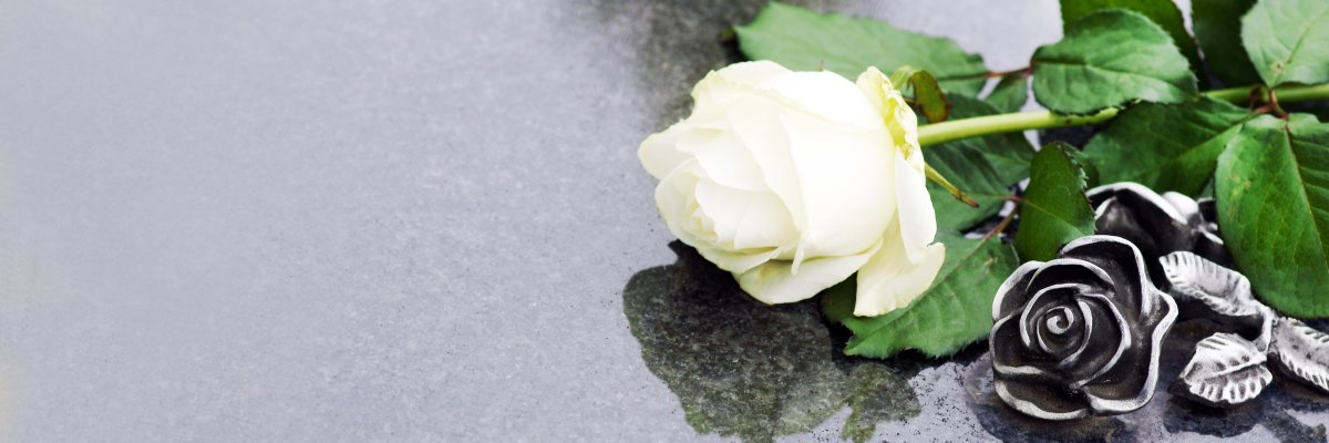 Auf einer glänzend spiegelnden Grabplatte liegt eine weiße Rose neben einer auf der Grabplatte angebrachten Rose aus Metall.