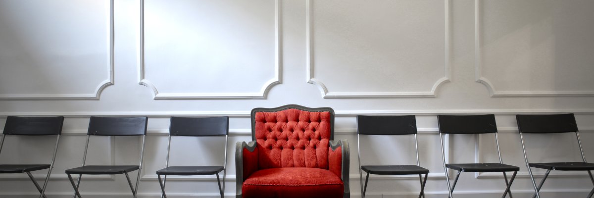 In einem Wartebereich mit Stuck verzierter Wand steht eine Reihe mit Klappstühlen. In der Mitte der Reihe steht ein großer, bequemer mit rotem Samt bezogener Sessel.