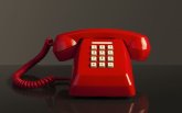 Vor einem dunklen Hintergrund steht ein rotes Telefon mit weißen Tasten. Auf dem Telefon liegt ein roter Hörer, der mit einem roten Kabel  mit dem Telefon verbunden ist.