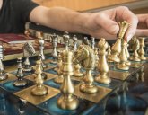 Schachspiel mit goldbarbigen und blauen Feldern und mit goldfarbigen Schachfiguren. Eine Hand hebt eine Schachfiguran, um einen Spielzug auszuführen.