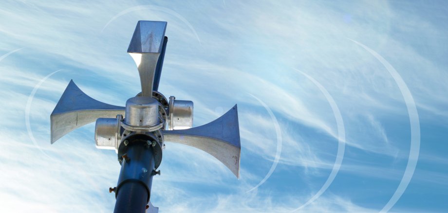 Metallmast mit Lautsprechern, die in alle vier Richtungen zeigen und eine Sirenenanlage darstellen, vor dem Hintergrund eines blauen Himmels. Weiße Halbkreise als Symbol für Schallwellen sind zu sehen.
