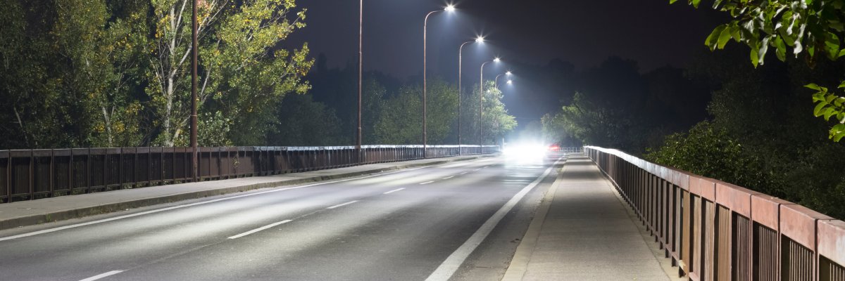 Autobrücke in der Nacht. Auf der linken Seite stehen Straßenlaternen in Reihe angeordnet. Auf der linken Fahrspur kommt ein Auto, welches den Betrachter blendet. Rechter Hand ist ein Brückengeländer. Das Brückengeländer ist rechts und links von Bäumen umsäumt.