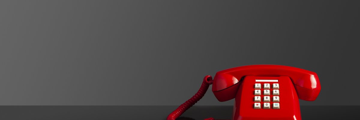 Vor einem dunklen Hintergrund steht ein rotes Telefon mit weißen Tasten. Auf dem Telefon liegt ein roter Hörer, der mit einem roten Kabel  mit dem Telefon verbunden ist.