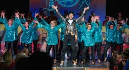 Mann mit Zylinder und goldenem Sakko vor 14 Frauen mit türkis glitzernden Oberteilen. Alle strecken die Arme nach oben. Tanzauftritt der TSG Blau Silber.