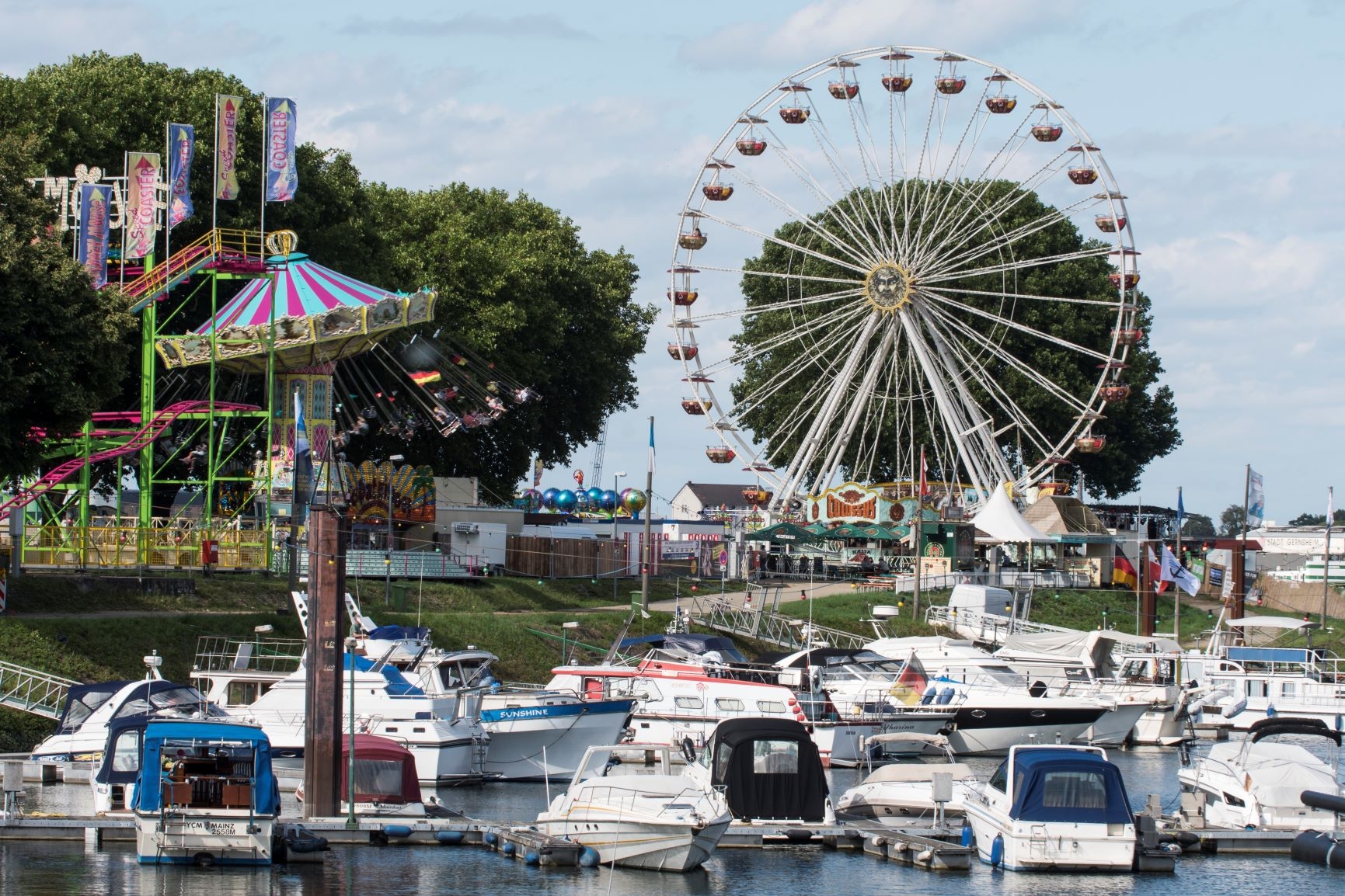 Hafenbecken während des mit Blick zur Hafenspitze. Im Hafenbecken liegen Sportboote. Auf dem Festplatz sieht man links das Kettenkarussell und rechts, im Hauptfokus, das Riesenrad. Überall wehen bunte Fahnen.