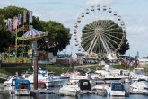 Hafenbecken während des mit Blick zur Hafenspitze. Im Hafenbecken liegen Sportboote. Auf dem Festplatz sieht man links das Kettenkarussell und rechts, im Hauptfokus, das Riesenrad. Überall wehen bunte Fahnen.
