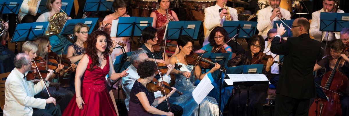 Neujahrskonzert in der Stadthalle mit den Frankfurter Philharmonikern. Die Solistin steht vor dem Orchester und trägt ein formelles burgundfarbenes Abendkleid. Rechts von ihr steht der Dirigent und blickt in ihre Richtung. Die Damen des Ensembles tragen alle Abendkleider die Herren Anzüge.