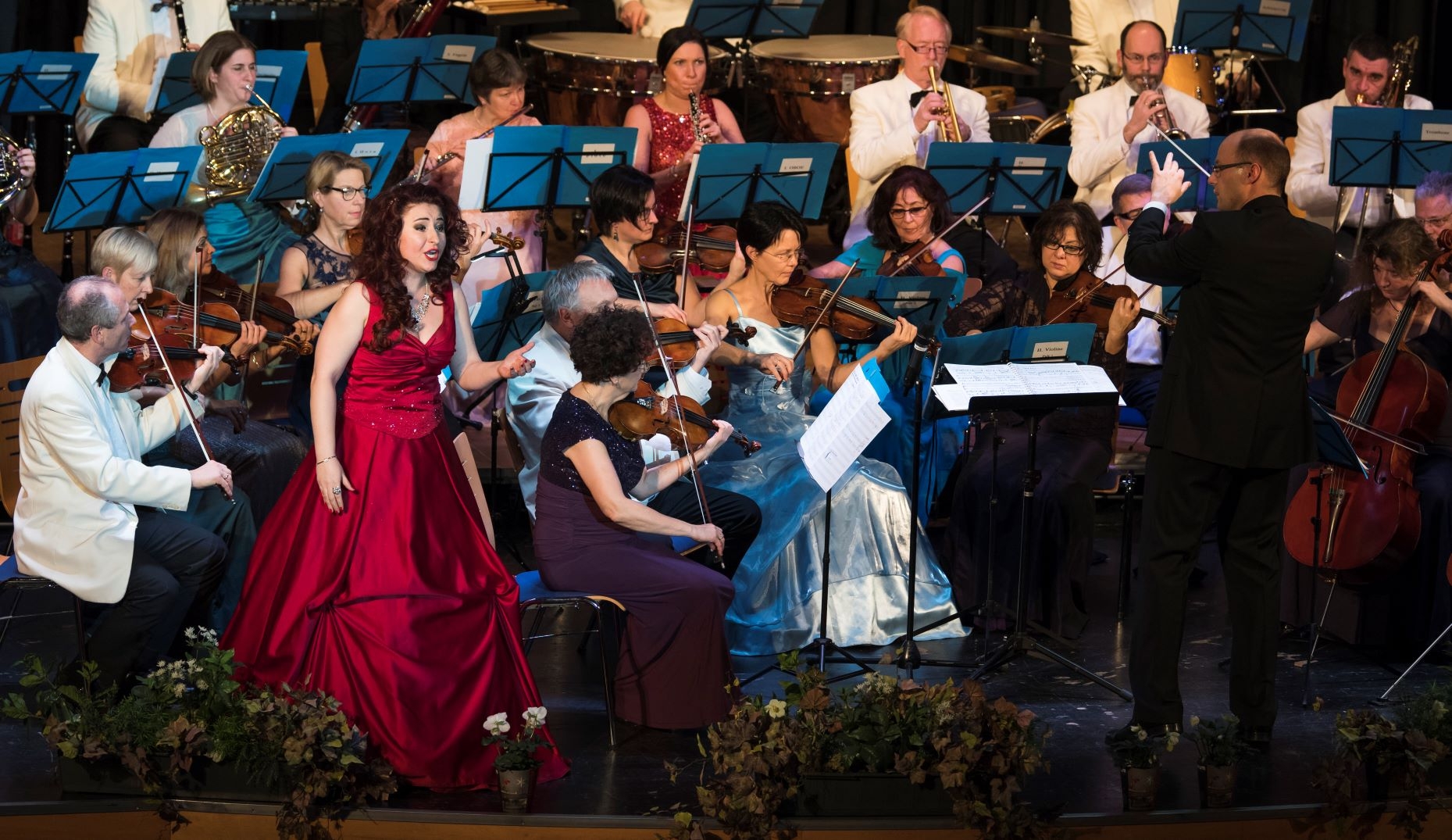 Neujahrskonzert in der Stadthalle mit den Frankfurter Philharmonikern. Die Solistin steht vor dem Orchester und trägt ein formelles burgundfarbenes Abendkleid. Rechts von ihr steht der Dirigent und blickt in ihre Richtung. Die Damen des Ensembles tragen alle Abendkleider die Herren Anzüge.