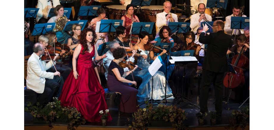 Neujahrskonzert in der Stadthalle mit den Frankfurter Philharmonikern. Die Solistin steht vor dem Orchester und trägt ein formelles burgundfarbenes Abendkleid. Rechts von ihr steht der Dirigent und blickt in ihre Richtung. Die Damen des Ensembles tragen alle Abendkleider die Herren Anzüge.