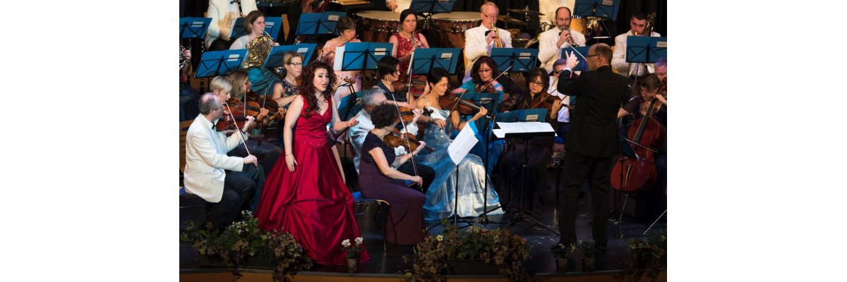 Neujahrskonzert in der Stadthalle mit den Frankfurter Philharmonikern. Die Solistin steht vor dem Orchester und trägt ein formelles burgundfarbenes Abendkleid. Rechts von ihr steht der Dirigent und blickt in ihre Richtung. Die Damen des Ensembles tragen alle Abendkleider die Herren Anzüge.
