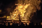 Pyramide aus Baumstämmen. Sie brennt lichterloh. Davor stehen mehrere Zuschauer.
