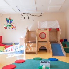 Der Gruppenraum zeigt eine Hochebene, die mit einer Rutsche ausgestattet ist. Links davon ist eine Kuschelecke. Im Vordergrund ist ein Teil eines Sitzkreises zu sehen.