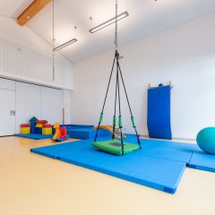 Im Turnraum sind Turnmattten auf dem Boden. Ein großer Gymnastikball liegt auf einer der Matten. Mittig hängt von der Decke eine große Korbschaukel.
