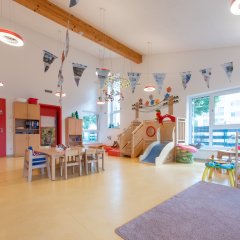 Es ist ein bunter Gruppenraum zu sehen. In ihm steht eine Tischgruppe mit Stühlen, eine Hochebene und rechts im Bild ist ein Regal mit Spielzeugen zu sehen.