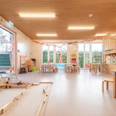 Der Gruppenraum ist hell und lichtdurchflutet. Links auf dem Boden sind Holzklötze und Schleichtiere  aufgebaut und Schleichtiere. Im Raum steht eine Tafel und mehrere Tischgruppen. Rechts ist eine Hochebene.