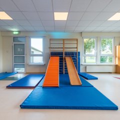 In der Raummitte sind mehrere Turnmatten auf dem Boden. An der Sprossenwand ist eine Bank eingeharkt, sodass man darauf runterrutschen kann.