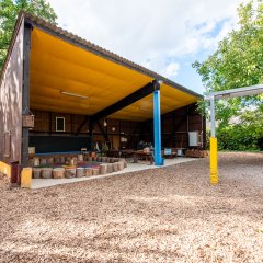 Das Bild zeigt die Basis 1. Das Carport ist zu sehen, in dem ein Stuhlkreis aus Stämmen und Tischgruppen stehen.