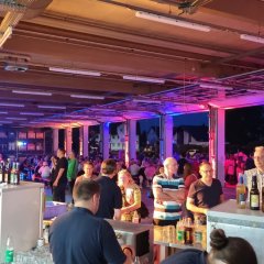 Rot und blau beleuchtete Fahrzeughalle der Feuerwehr. Die Fahrzeuge wurden herausgefahren. Vor und in der Halle sind Biergartengarnituren aufgestellt an denen Menschen sitzen. In der ganzen Halle stehen eine Vielzahl von Menschen. Im Vordergrund eine Theke mit Zapfanlage.