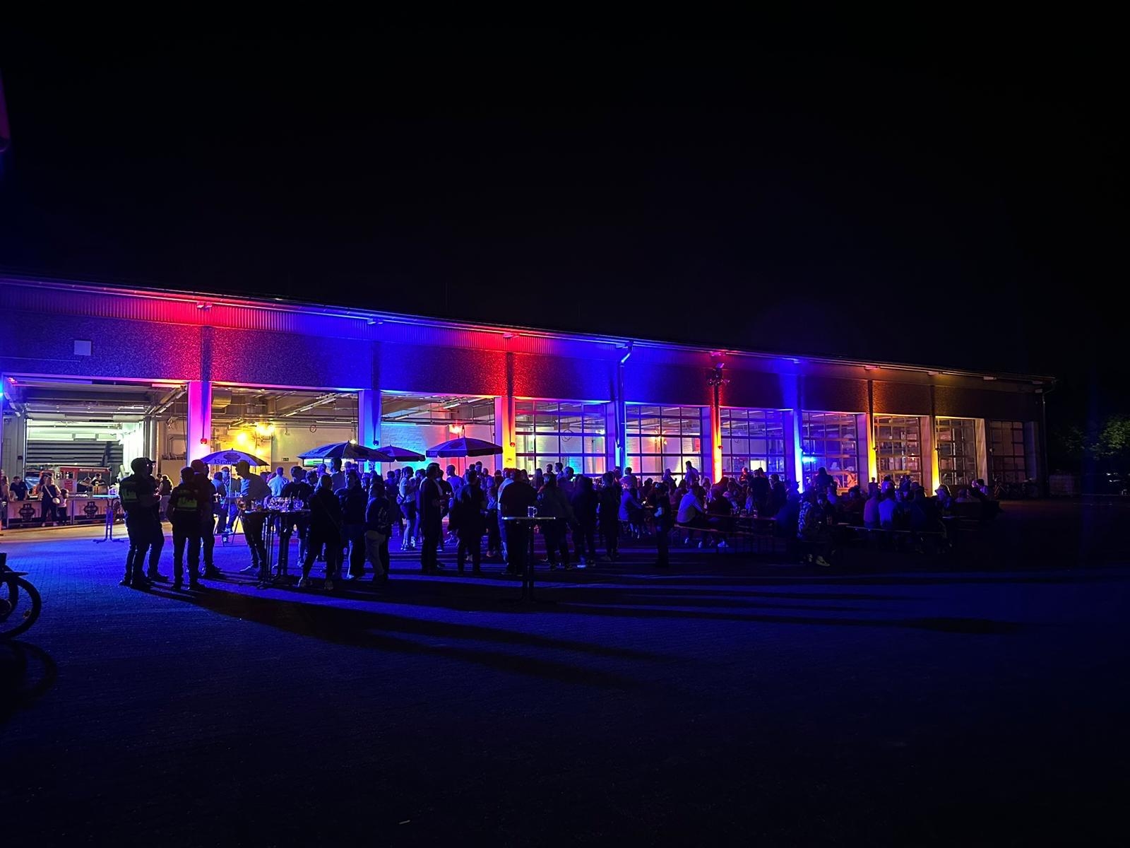 Blick von außen auf die rot und blau beleuchtete Fahrzeughalle der Feuerwehr. Die Fahrzeuge wurden herausgefahren. Vor und in der Halle sind Biergartengarnituren aufgestellt an denen Menschen sitzen. Vor und in der Halle stehen eine Vielzahl von Menschen.