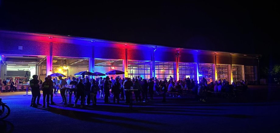 Blick von außen auf die rot und blau beleuchtete Fahrzeughalle der Feuerwehr. Die Fahrzeuge wurden herausgefahren. Vor und in der Halle sind Biergartengarnituren aufgestellt an denen Menschen sitzen. Vor und in der Halle stehen eine Vielzahl von Menschen.