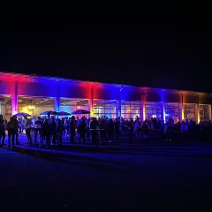 Blick von außen auf die rot und blau beleuchtete Fahrzeughalle der Feuerwehr. Die Fahrzeuge wurden herausgefahren. Vor und in der Halle sind Biergartengarnituren aufgestellt an denen Menschen sitzen. Vor und in der Halle stehen eine Vielzahl von Menschen.