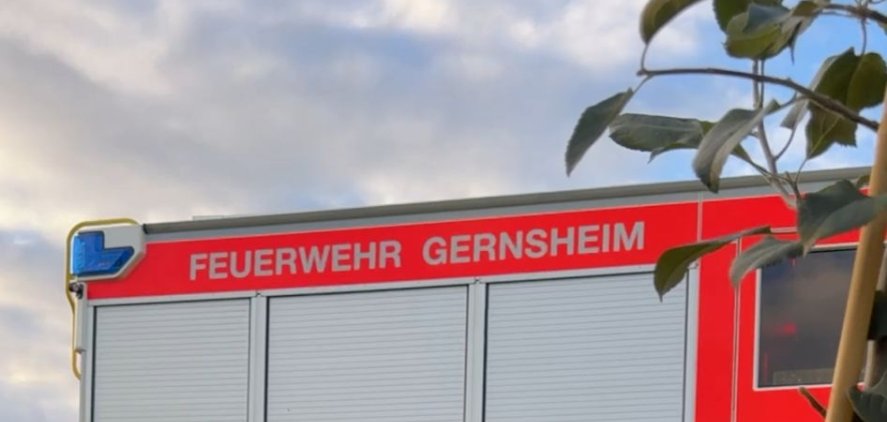 Eine behandschuhte Hand klopft die Erde um einen frisch gesetzten Apfelbaum fest. Im Hintergrund ein Feuerwehrfahrzeug.