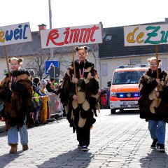 Drei Kinder in Eulenkostümen laufen auf der Straße. Dahinter ein Feuerwehrauto. Die Straße ist beidseitig von Menschen gesäumt.